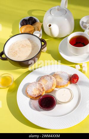 Assapora una deliziosa colazione con frittelle di formaggio, farinata d'avena cremosa, tè aromatico, miele dorato, marmellata, e frutta fresca su un tavolo giallo soleggiato, c Foto Stock