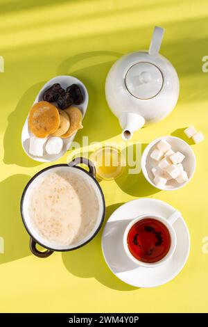 Una colazione accogliente con farinata d'avena con latte, tè in una tazza bianca su piattino, miele in una ciotola di vetro e fette di pane su sfondo giallo. Shad Foto Stock