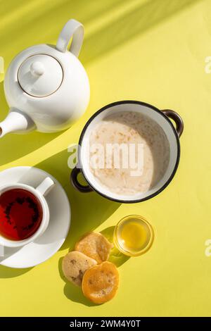 Una colazione accogliente con farinata d'avena con latte, tè in una tazza bianca su piattino, miele in una ciotola di vetro e fette di pane su sfondo giallo. Shad Foto Stock