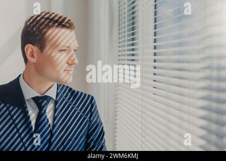 Il giovane uomo d'affari guarda con attenzione attraverso le tende veneziane, bagnate dalla luce del sole. Foto Stock