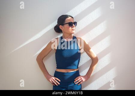 donna sportiva in piedi contro un muro al sole, con occhiali da sole. Foto Stock