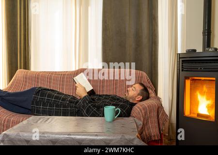 Uomo ispanico con la barba di 40 anni sdraiato sul divano a leggere un libro, è molto a suo agio con il calore di una stufa a pellet Foto Stock