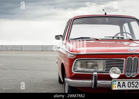 BMW rossa classica degli anni '1960 L'angolo anteriore della berlina tedesca è parcheggiato in un grande parcheggio di fronte al cielo nuvoloso. Foto Stock
