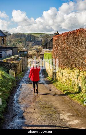 Donna con un cappotto rosso che cammina attraverso il villaggio di Eyam nel Derbyshire English Peak District durante l'inverno Foto Stock