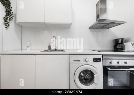 Immagine frontale di una piccola cucina arredata con mobili bianchi, ripiano bianco, con elettrodomestici bianchi e in acciaio inossidabile Foto Stock