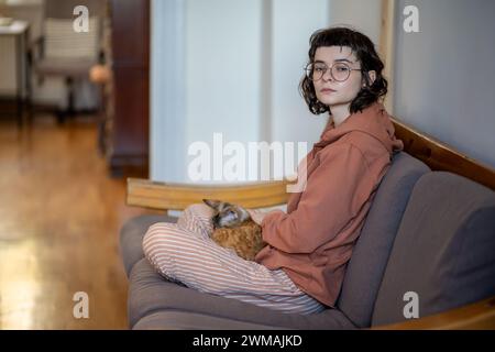 Una teenager stanca che accarezza il gatto che fa una pausa guardando il lato seduto sul divano di casa. Foto Stock