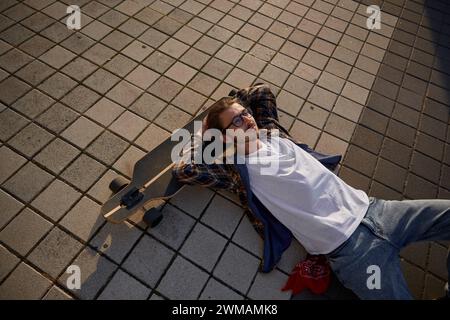 Ragazzo hipster disteso sullo skateboard dopo l'allenamento, rilassandosi al tramonto Foto Stock