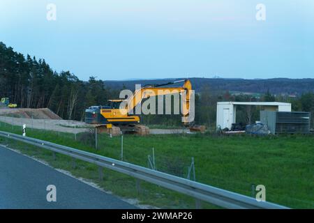 La scena cattura un escavatore giallo parcheggiato accanto a un mucchio di terra sul ciglio della strada, con recinzioni metalliche e una piccola struttura nelle vicinanze, adagiata contro un tr Foto Stock