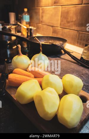 Verdure sbucciate su un asse di legno con un coltello. Patate crude, cipolla e carote sul tavolo da cucina. Preparare il concetto di cibo. Verdure sul tagliere. Foto Stock