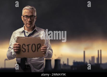 Un uomo d'affari sporco e disperato, con un cartello e protesta contro l'inquinamento, gli impianti industriali e i gas tossici sullo sfondo Foto Stock