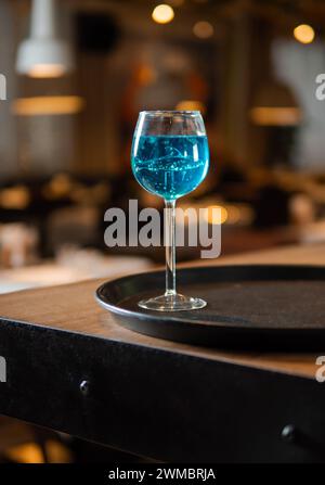 Bicchiere di cocktail laguna blu in un bicchiere da cocktail a gambo alto di squalo sullo sfondo del bellissimo bancone del bar con bokeh. Foto estetica del cocktail laguna blu alla luce della sera, all'interno del ristorante o del bar Foto Stock