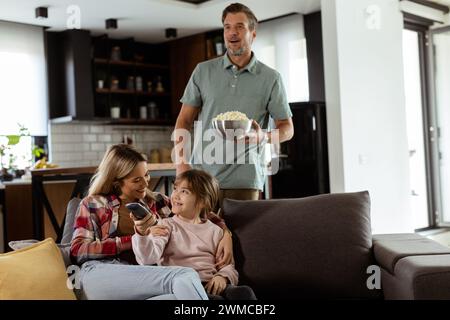 Una famiglia di tre persone si trova comodamente adagiata su un divano, con i volti che riflettono l'emozione e l'attenzione mentre condividono una ciotola di popcorn durante una sosp Foto Stock