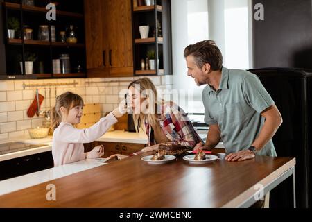 Una scena emozionante si svolge mentre una famiglia assaggia insieme una deliziosa torta al cioccolato nel calore della sua cucina soleggiata, condividendo sorrisi e sorrisi Foto Stock