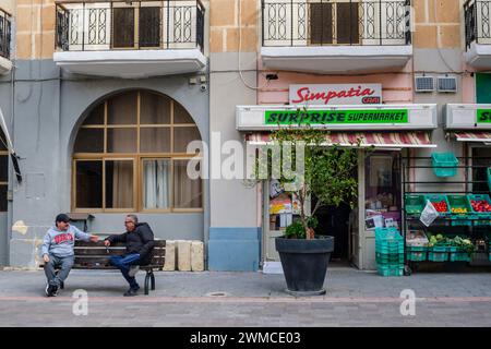 Due uomini che chiacchierano su una panchina fuori da un negozio di alimentari locale a Senglea, la Valletta, Malta Foto Stock