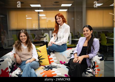 Tre donne professionali sedute casualmente su un vivace divano a motivi geometrici in un ufficio creativo. Foto Stock