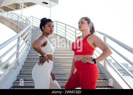 Due donne più grandi e sicure di sé in abbigliamento fitness stanno insieme su una scala all'aperto. Femmine con lo stesso tipo di corpo che posano all'aperto dopo il fitness Foto Stock