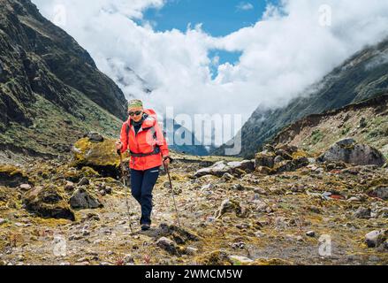Donna con occhiali da sole con zaino e bastoncini da trekking vestita di giacca softshell rossa durante il trekking nel Parco Nazionale Makalu Barun in Nepal. Escursione in montagna Foto Stock