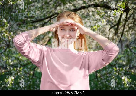 la giovane ragazza dai capelli rossi sorride in primavera, guarda con le mani chiuse gli occhi dal sole Foto Stock