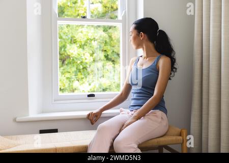 La giovane donna birazziale si siede contemplativamente vicino a una finestra a casa Foto Stock