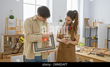 Un uomo e una donna in una luminosa officina esaminano insieme una cornice vintage, tra utensili per la lavorazione del legno e legno. Foto Stock