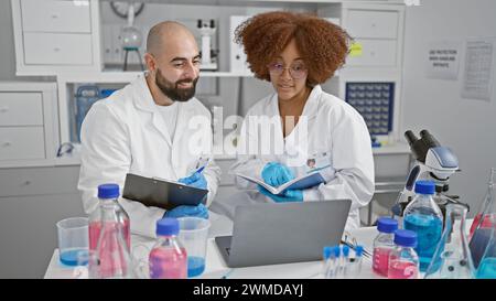 Due scienziati sorridenti si impegnano in una conferenza online, leggendo un libro e scrivendo sugli appunti nel loro laboratorio mentre lavorano a una ricerca medica Foto Stock