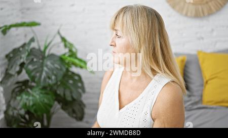 Bella donna bionda matura con un'espressione seria che guarda di lato, rilassandosi comodamente nel letto, all'interno di una camera da letto Foto Stock
