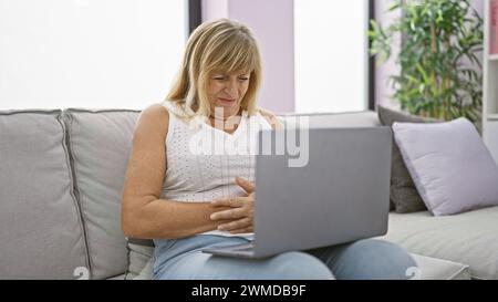 Il pancino doloroso trasforma il laptop di una donna bionda di mezza età in un'estenuante calvario a casa. Foto Stock