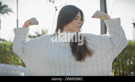 Donna ispanica che si flette con sicurezza in un parco con vegetazione e indossa un maglione bianco. Foto Stock