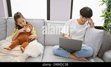 Madre-figlia che si lega a casa, donna che lavora mentre adorabile ragazza si siede sul divano abbracciando la bambola scimmia, abbracciando uno stile di vita informale Foto Stock