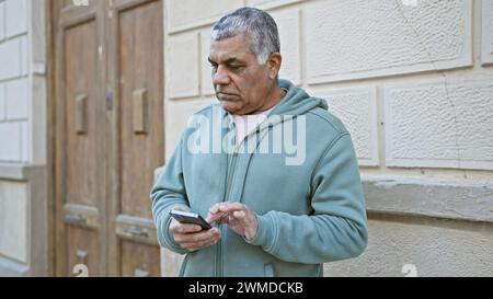 Un uomo dai capelli grigi maturo con cappuccio usa lo smartphone per le strade della città Foto Stock