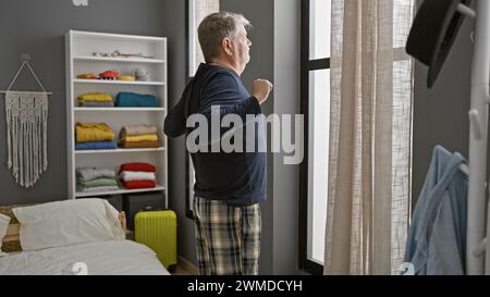Un uomo dai capelli grigi in pigiama si allunga mentre guarda fuori dalla finestra in una camera da letto ordinata. Foto Stock
