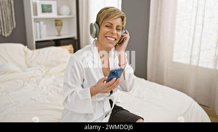 Donna sorridente con capelli corti che ascolta la musica con le cuffie e tiene lo smartphone in una camera da letto luminosa Foto Stock