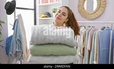 Una giovane donna ispanica tiene una pila di maglioni piegati in una stanza dei vestiti con uno specchio del guardaroba. Foto Stock