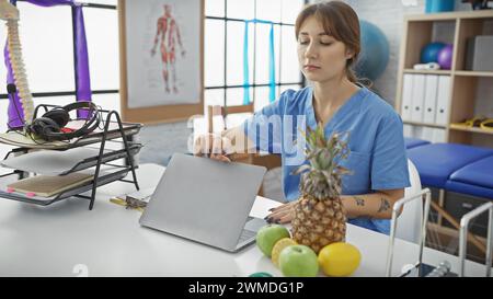 Una donna caucasica concentrata in scrub blu che lavora su un laptop in una brillante clinica di fisioterapia con attrezzature per l'esercizio fisico Foto Stock