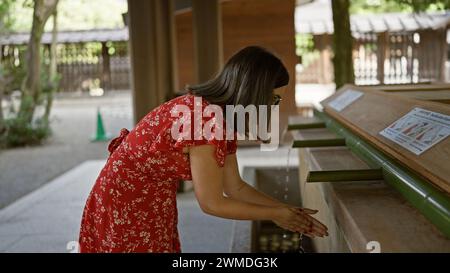 La bella donna ispanica con gli occhiali partecipa al tradizionale rituale purificante asiatico al tempio meiji, lavandosi le mani in acqua naturale fluente da Foto Stock