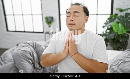 Mattinata piena di fede, giovane e bello uomo cinese che esprime la sua fede in gesù attraverso la preghiera mentre si rilassa su un comodo letto nell'accogliente interi Foto Stock