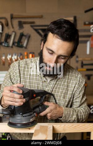 Foto verticale un abile falegname caucasico medio adulto leviga con cura la gamba di una sedia di legno, dimostrando attenzione ai dettagli in un'officina ordinata Foto Stock