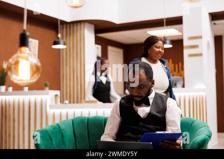 Responsabile del settore alberghiero afroamericano che guarda lo schermo del computer mentre si siede nella hall dell'hotel per gestire le forniture. Un fattorino nero che usa un computer portatile per organizzare il trasporto Foto Stock