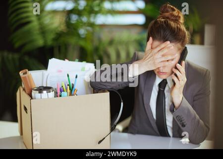 Nuovo lavoro. lavoratrice donna moderna e triste in un ufficio verde moderno in un abito da lavoro grigio con effetti personali in una scatola di cartone che parla su uno smartphone. Foto Stock