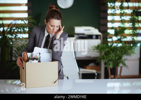 Nuovo lavoro. lavoratrice moderna preoccupata in un moderno ufficio verde in abito da lavoro grigio con effetti personali in scatola di cartone. Foto Stock