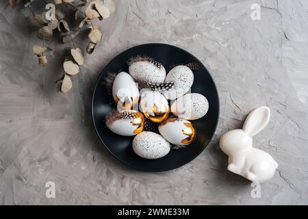 Piatto nero con uova di Pasqua, foglie di eucalipto e coniglietto su sfondo grigio testurizzato. Vista dall'alto, disposizione piatta. Foto Stock