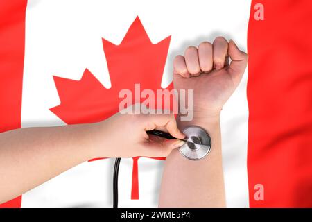 La testa di uno stetoscopio medico sul polso (primo piano) sullo sfondo della bandiera canadese. Il concetto di sistema sanitario canadese Foto Stock