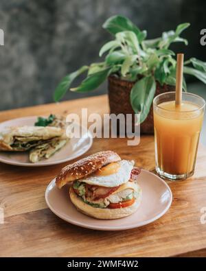 Primo piano di un sandwich con bagel per colazione con uova strapazzate, spinaci, formaggio, pomodoro. bevanda di vetro e pianta Foto Stock