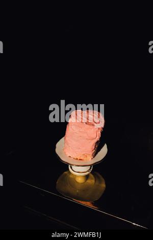 piccola torta a forma di cuore alta e pendente con glassa rosa pallido su piedistallo in ceramica bianca su un tavolo dorato con sfondo nero Foto Stock