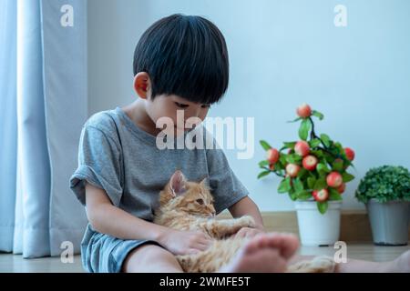 Un piccolo ragazzo asiatico tiene amorevolmente un gattino arancione. Foto Stock