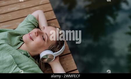 Donna rilassata indossando cuffie ascoltare musica seduto su un molo vicino al lago naturale in estate Foto Stock