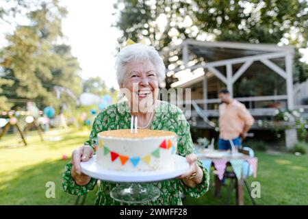 Bella donna anziana di compleanno con corona di carta che tiene la torta di compleanno. Festa di compleanno in giardino per una donna anziana. Foto Stock