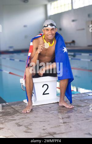 Un giovane nuotatore maschile birazziale festeggia a bordo piscina, drappeggiato da una bandiera australiana con una medaglia Foto Stock