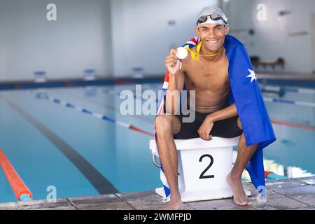 Il giovane nuotatore maschile birazziale festeggia a bordo piscina, drappeggiato da una bandiera australiana con una medaglia Foto Stock