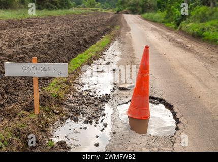 Poppa, profonda gola piena di acqua piovana, con cono stradale arancione e cartello a sinistra che indica la poppa sulla strada rurale a binario unico in Oriente Foto Stock